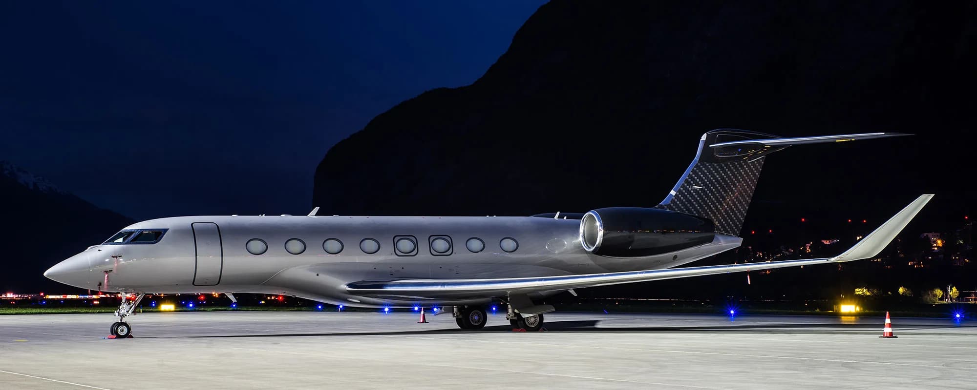 A silver and black Gulfstream G650 business jet parked on an airport apron at night with mountains in the background.