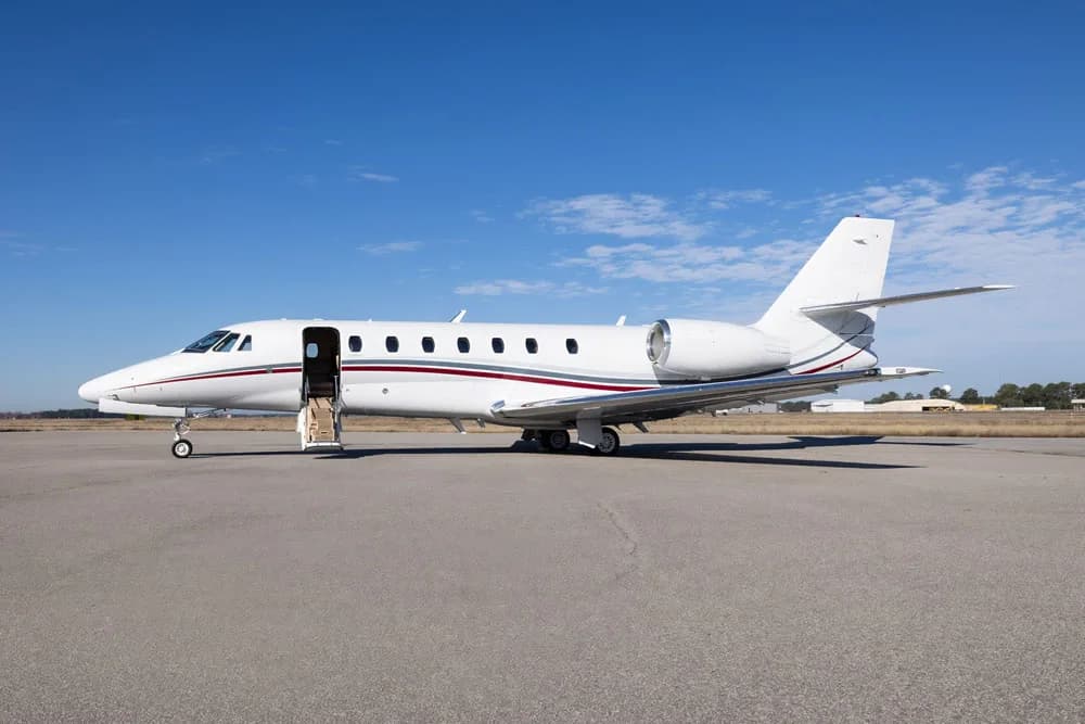A three-quarter front view of a white business jet with red and grey stripes on an airfield.