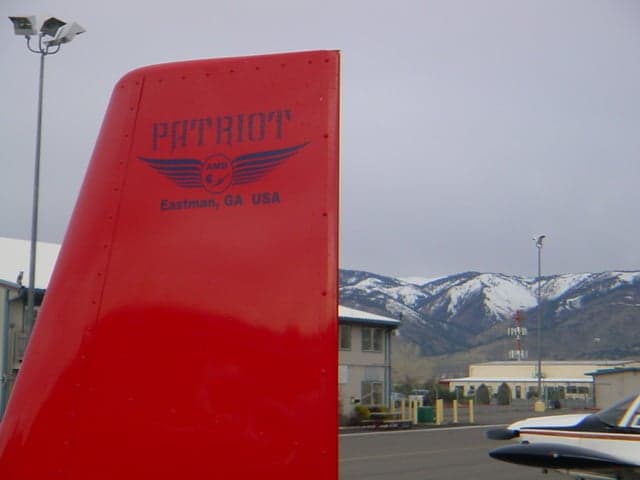 A close-up of the red tail of a Patriot aircraft, showing the manufacturer's logo, with snowy mountains in the background.
