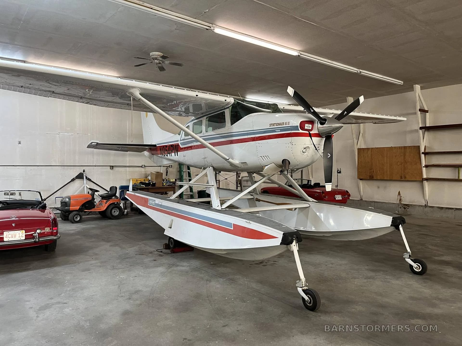 A white Cessna Stationair 6 II floatplane with red and blue stripes, registration N954PW, viewed from the front-right in a hangar.