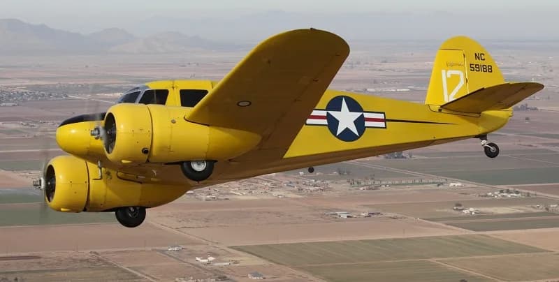 A yellow Beechcraft Model 18 flying over a rural landscape.