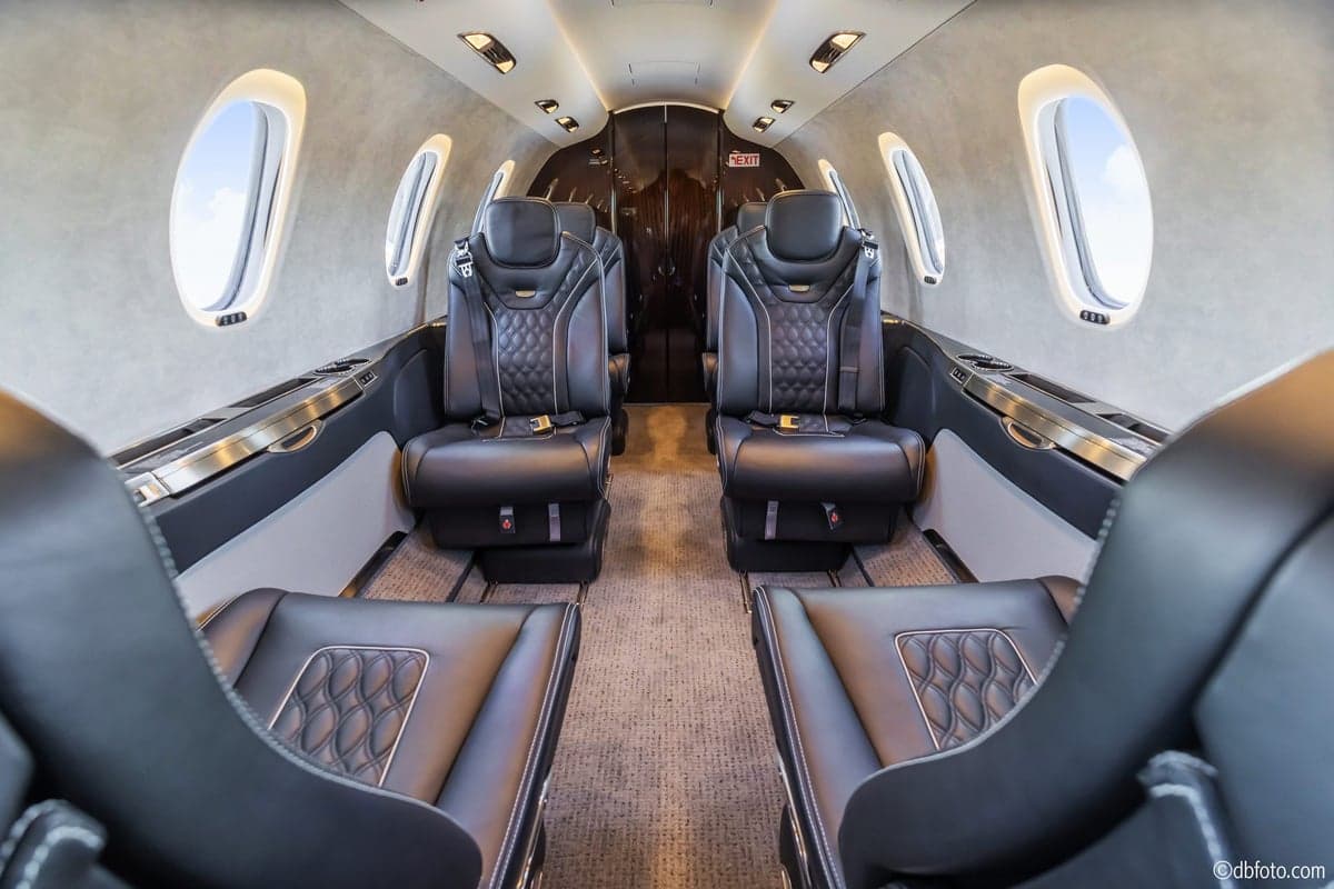 A wide view of a luxurious aircraft cabin with four black leather seats, quilted stitching, and large oval windows on both sides.