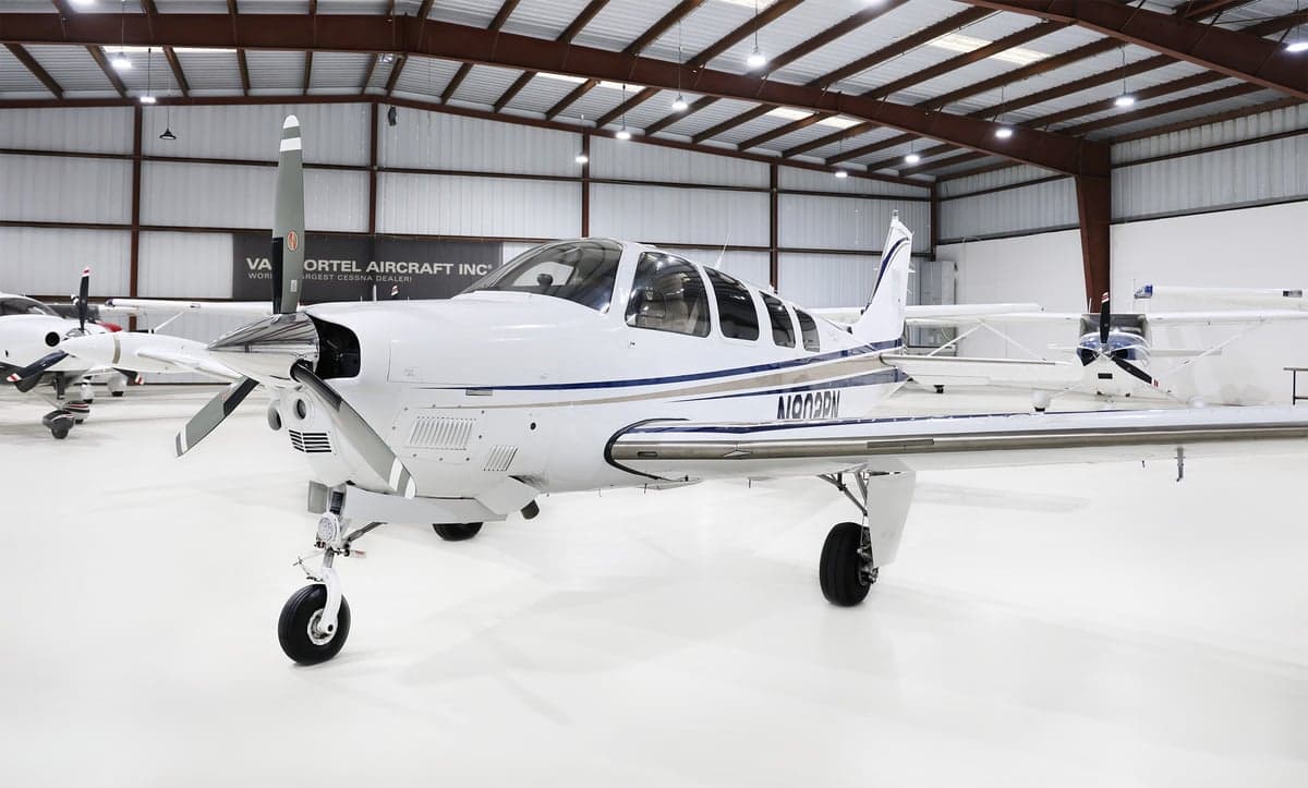 Wide front-side view of a white Beechcraft Bonanza N802PN with blue and grey stripes, parked in a hangar.