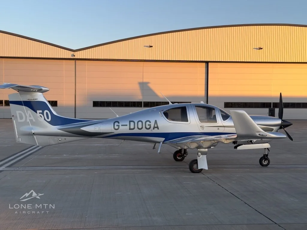 A side view of a silver and blue Diamond DA50 aircraft with registration G-DOGA parked on an airfield at sunset.