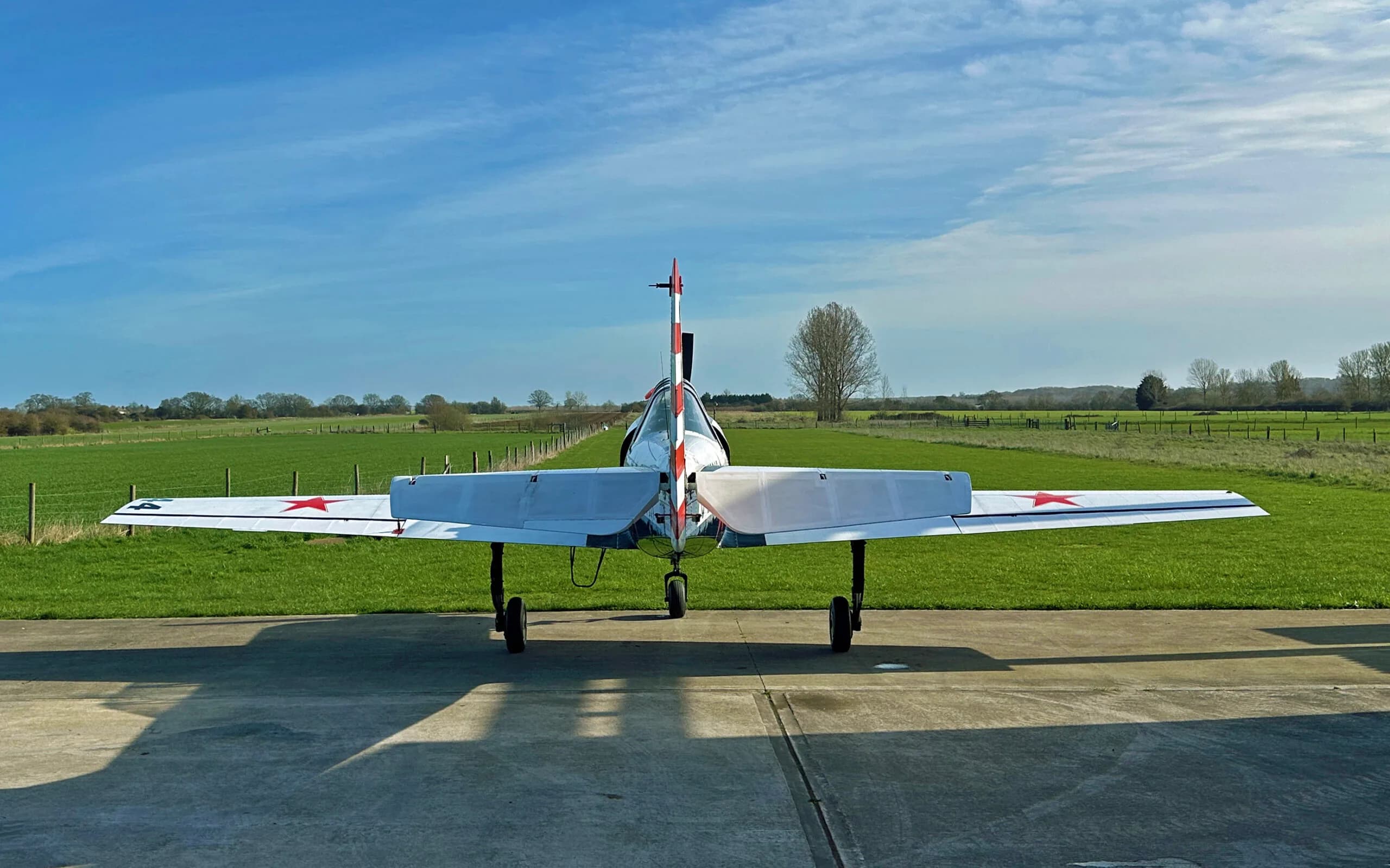A rear view of a white and black Yak-52 aircraft with red stars, parked on a concrete pad facing a grassy field.