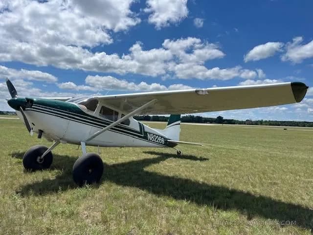 A side-front view of a white and green single-engine propeller aircraft with large tires, registration N82266, parked on a grassy field.