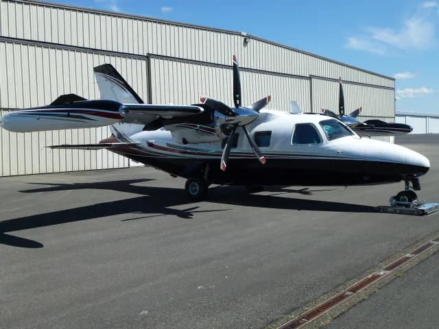 A white and black Mitsubishi MU-2 twin-turboprop aircraft with red and grey stripes, parked on an asphalt ramp in front of a hangar.