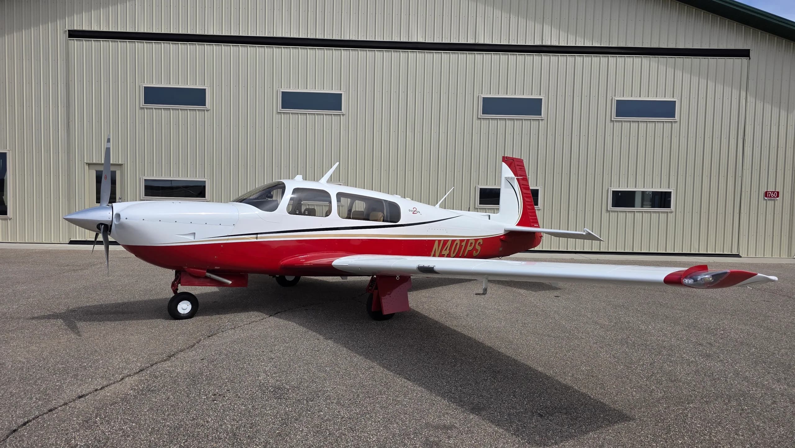 A full side view of a red and white Mooney aircraft with registration N401PS parked on an asphalt apron in front of a hangar.