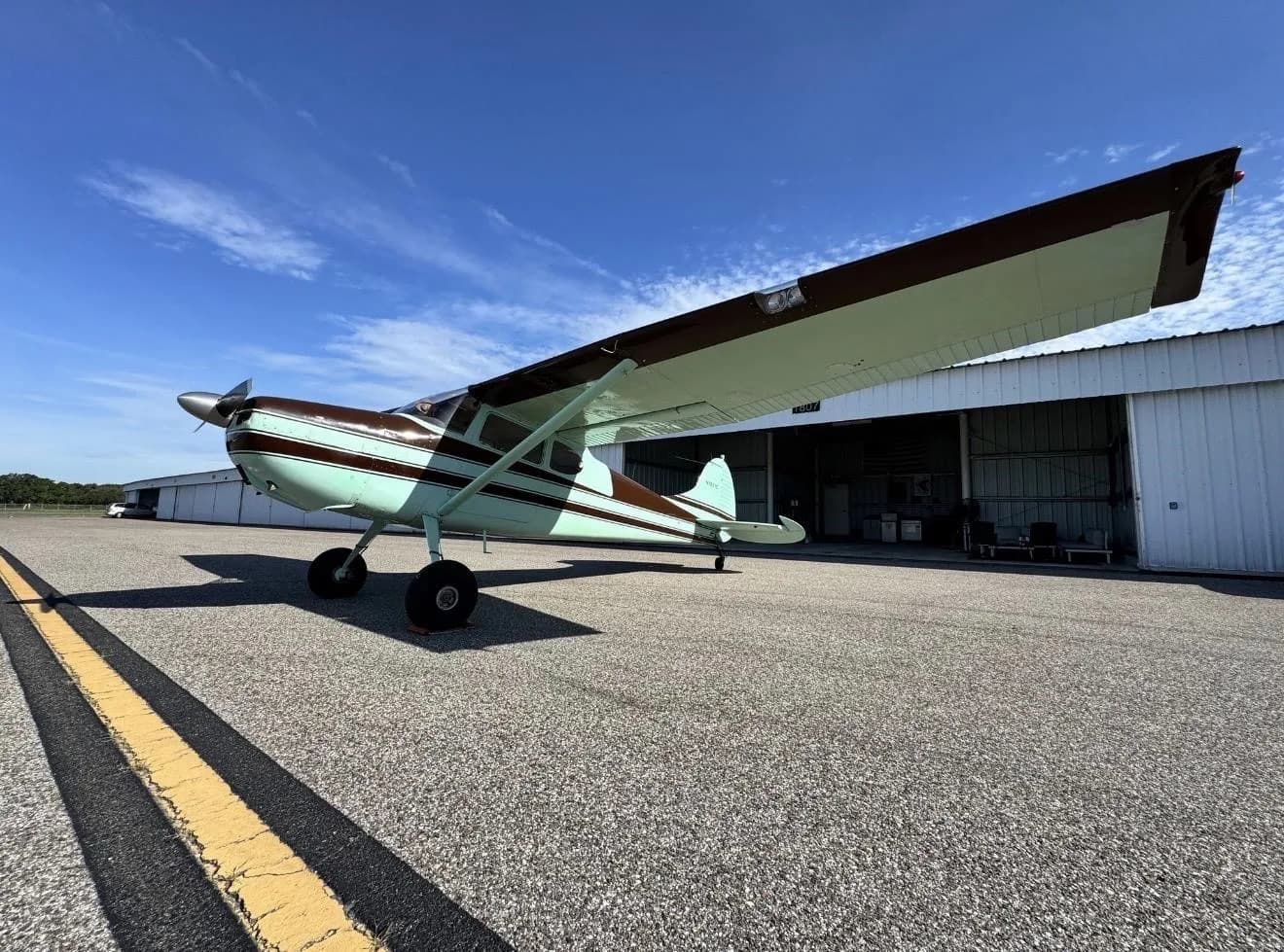 A brown and light green single-engine propeller aircraft with high wings is parked on an asphalt tarmac in front of hangars under a blue sky.