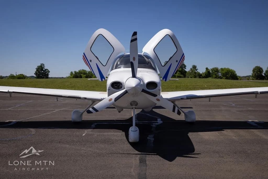 Front view of a white Cirrus aircraft (N218DP) with blue and red stripes, doors open, parked on an airfield.