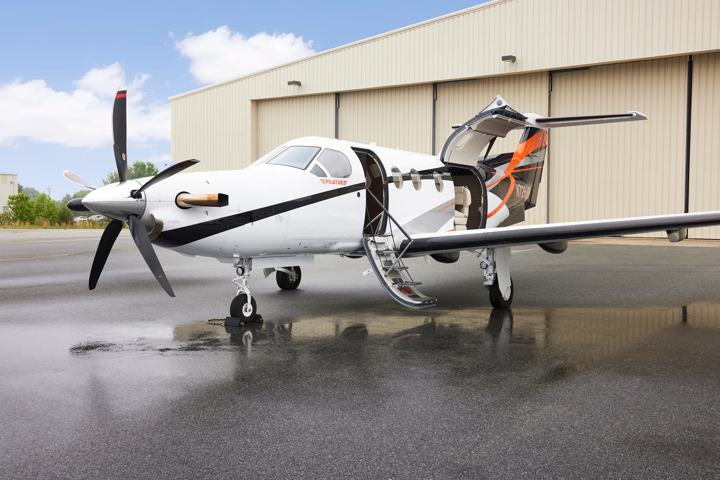 A white Pilatus PC-12 aircraft with black and orange accents and an open cabin door, parked on a wet tarmac in front of a hangar.