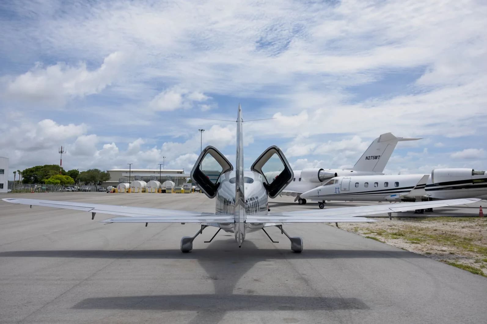 A rear view of the silver Cirrus SR22 aircraft with its gull-wing doors open, parked on an tarmac with other aircraft in the background.