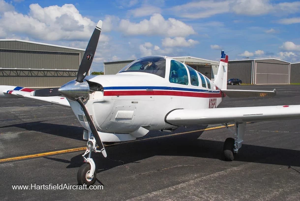 A white Beechcraft Bonanza aircraft with red and blue stripes is parked on an airfield tarmac with hangars in the background under a cloudy sky.