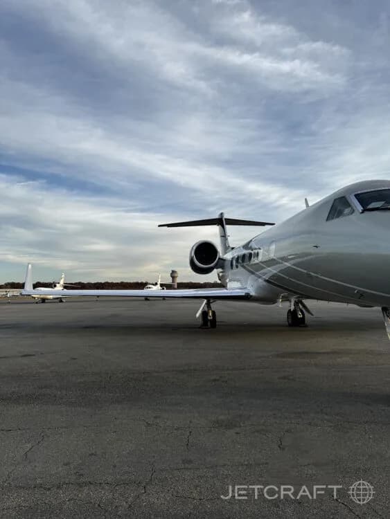 A close-up view of the silver and dark blue Gulfstream G450's engine and wing on a tarmac, with other aircraft and a control tower in the background.