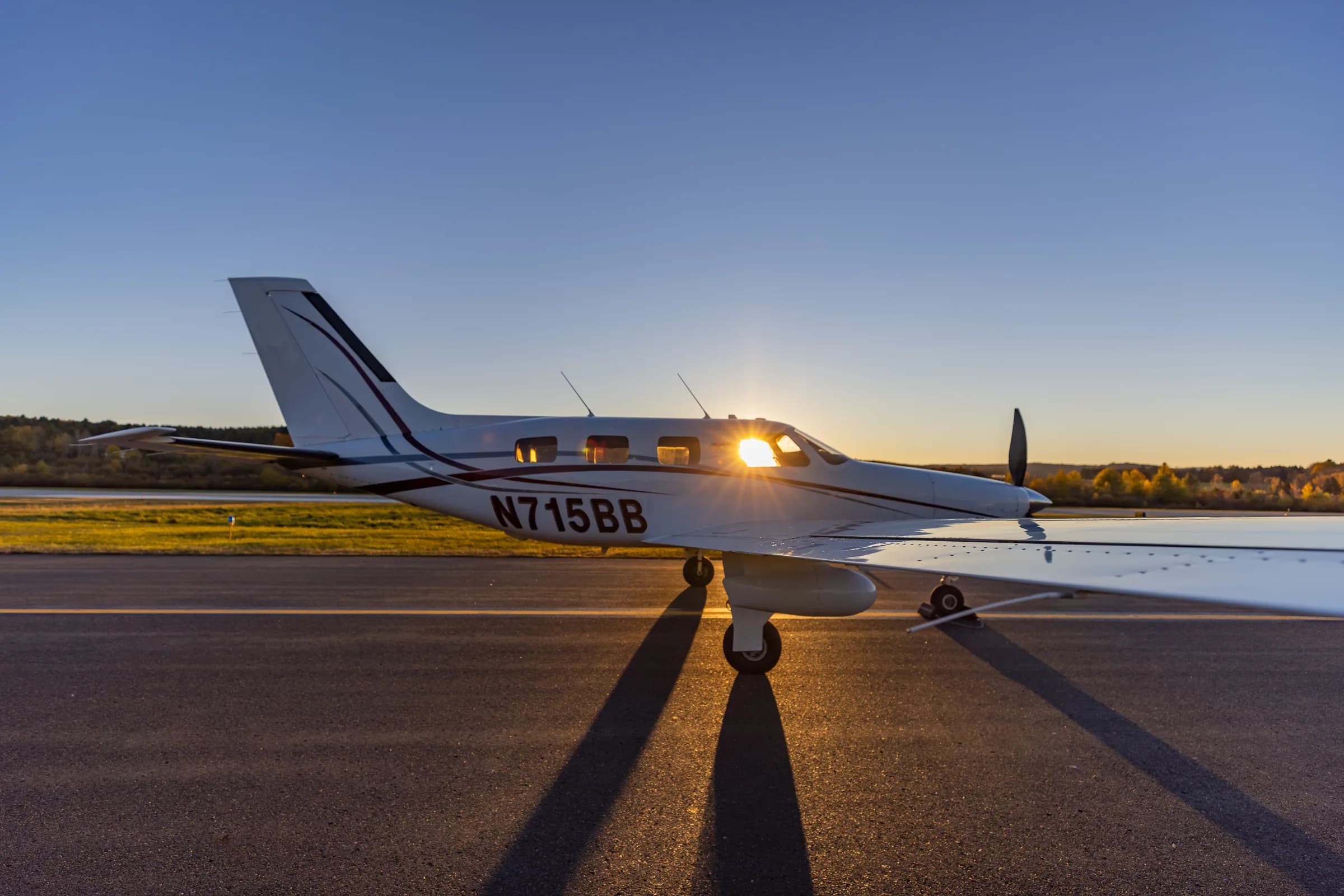 A side view of the Piper Malibu Mirage N715BB with the sun glinting through its windows on a runway.