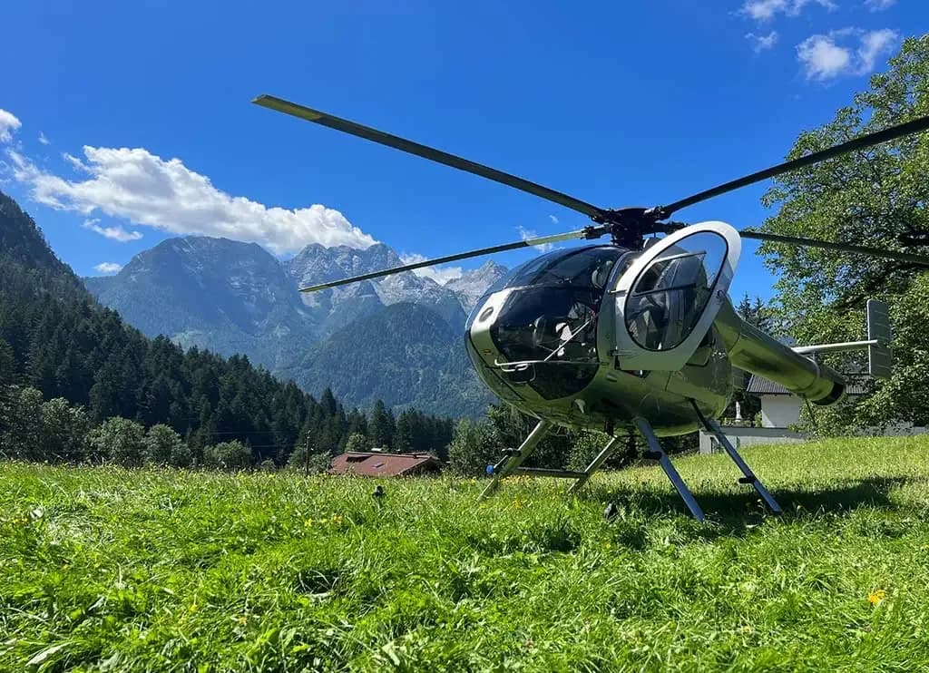 A silver helicopter is parked in a vibrant green grassy field with a clear blue sky and mountains in the background.