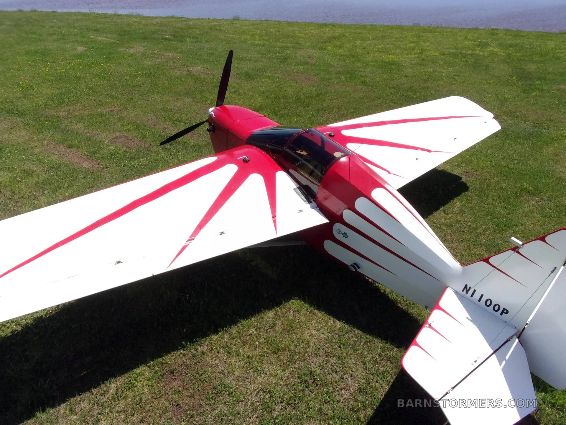 An aerial view of a red and white single-engine aircraft with a flame-like paint scheme, parked on grass near a body of water.
