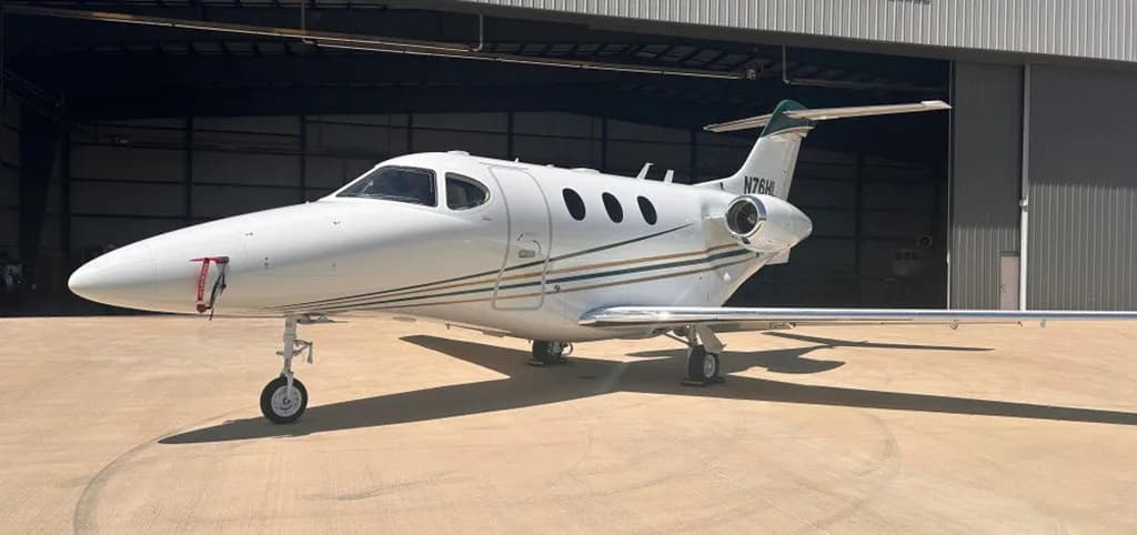 A white Beechcraft Premier 1 private jet, registration N76HL, with green and gold stripes, parked on a concrete tarmac in front of an open hangar.