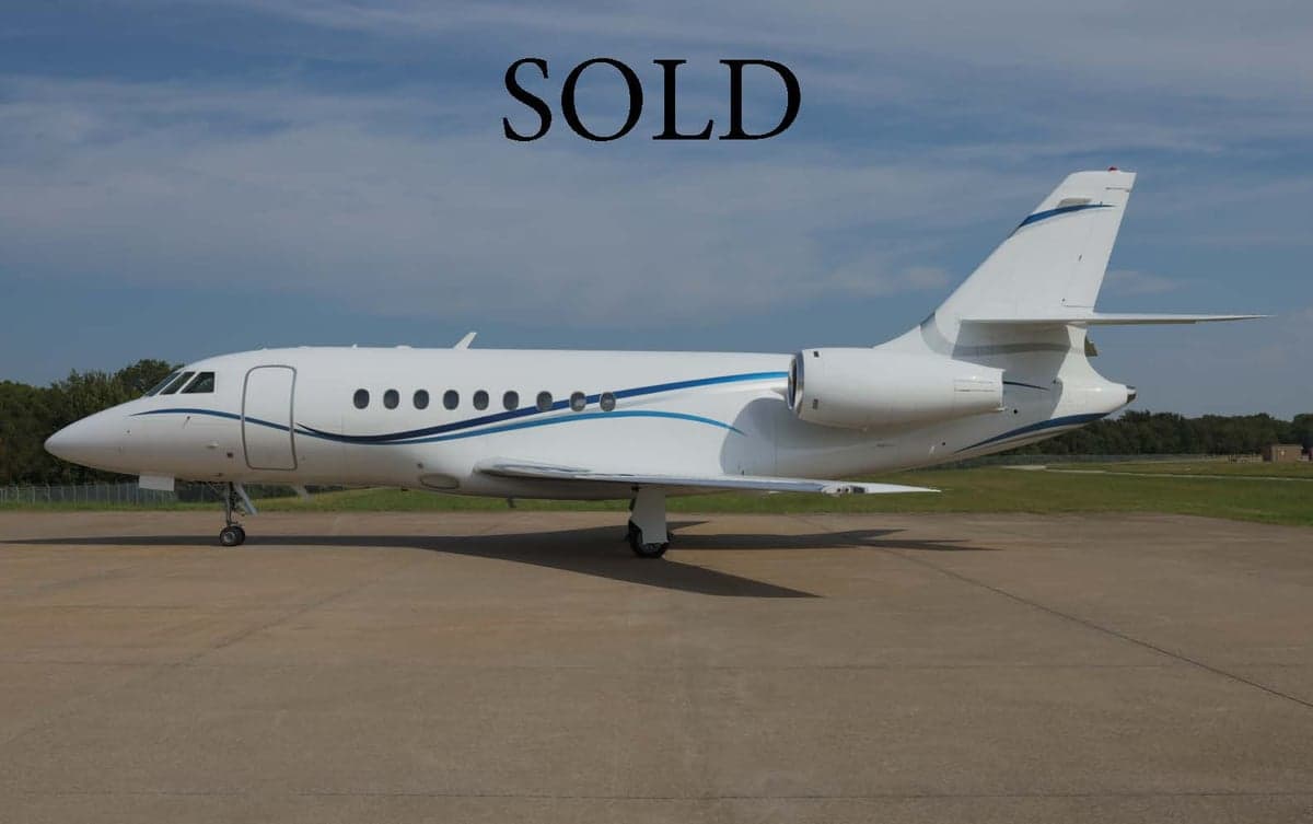 Side view of a white and blue Dassault Falcon jet on the tarmac with SOLD overlaid.