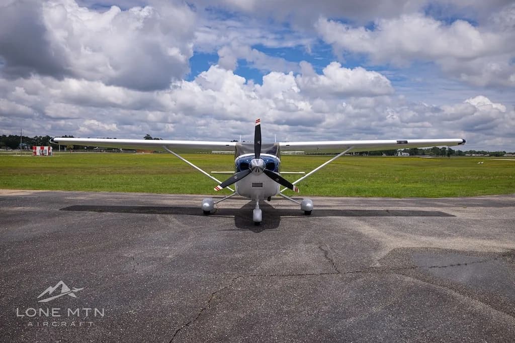 A head-on view of a white and blue Cessna Skylane TC (N6020Z) parked on an airfield under a partly cloudy sky.