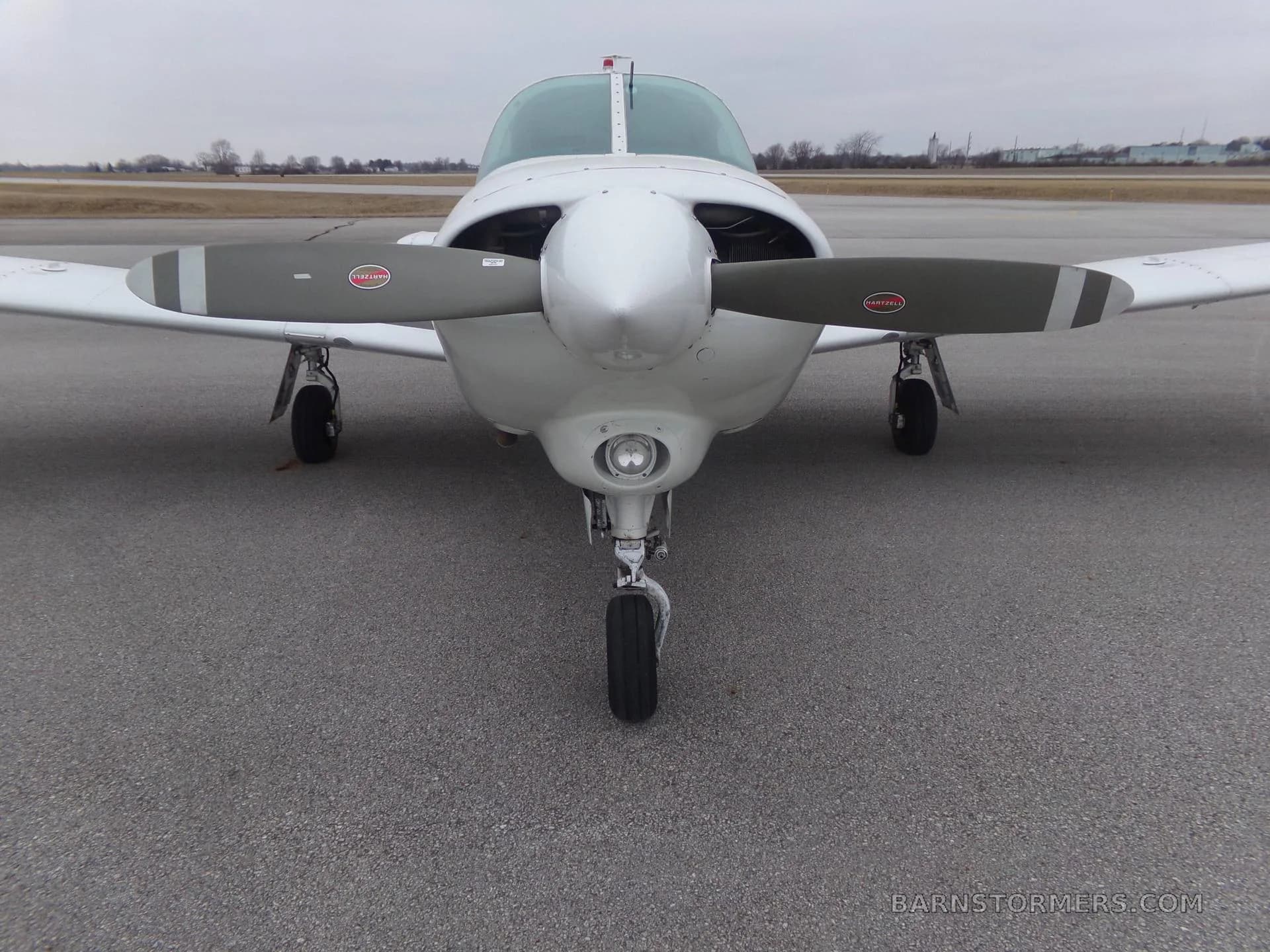 A white Piper Turbo Arrow III aircraft is viewed head-on from the front, showing its propeller and landing gear on an asphalt tarmac.