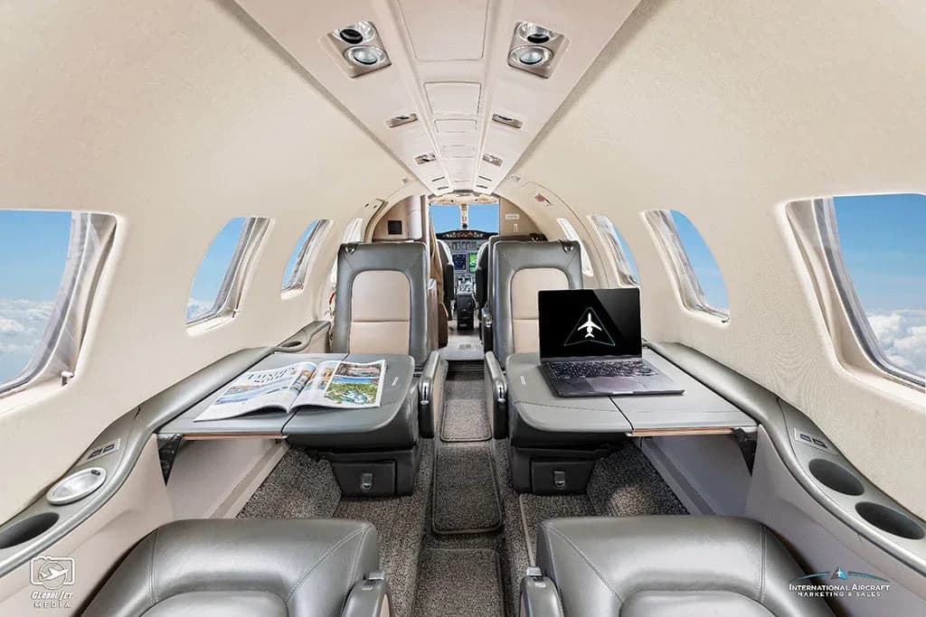 The interior cabin of a business jet shows grey and tan leather seats with tables extended, a laptop, and a magazine.