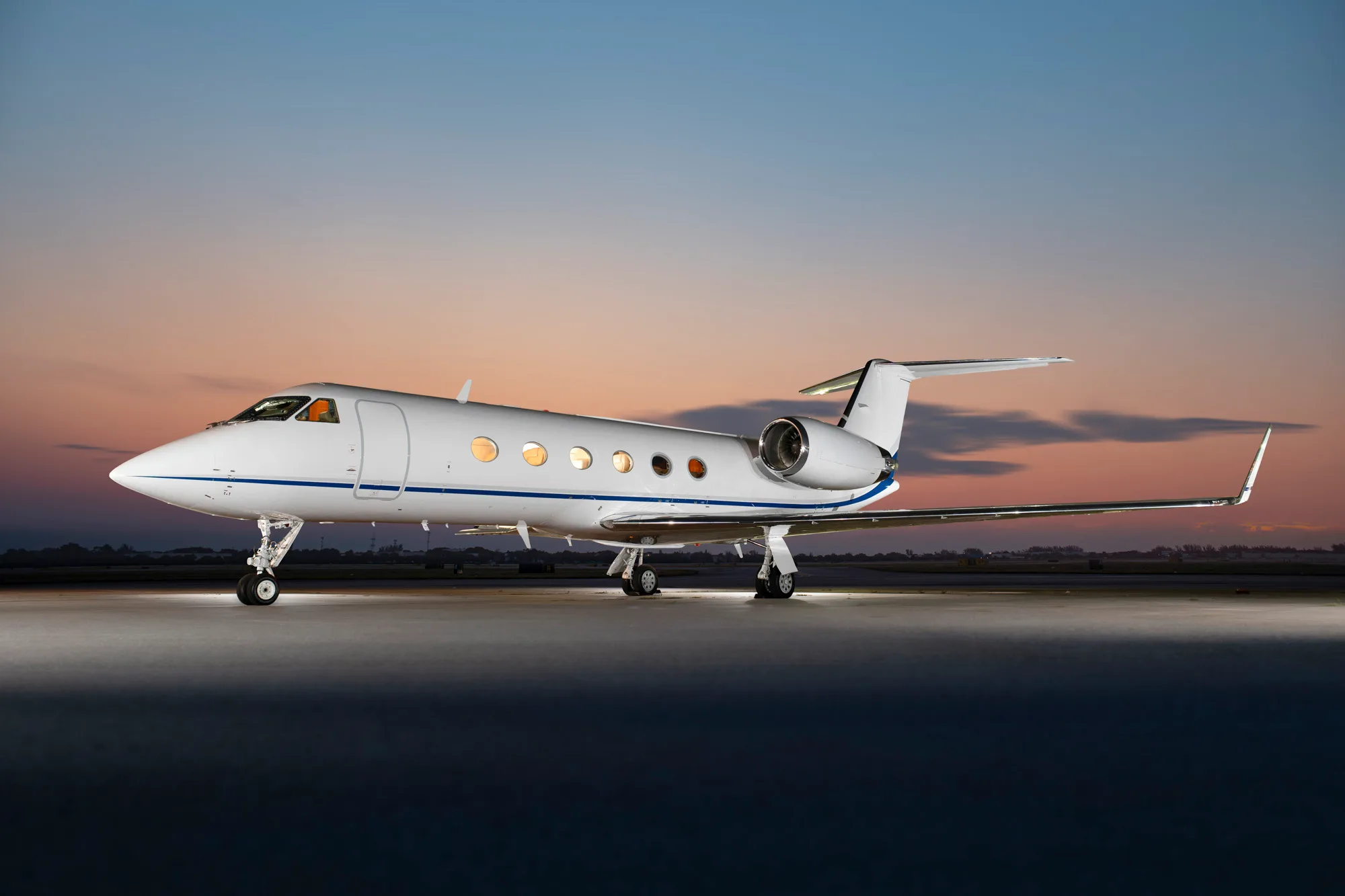 A white business jet with a blue stripe parked on a tarmac at sunset.