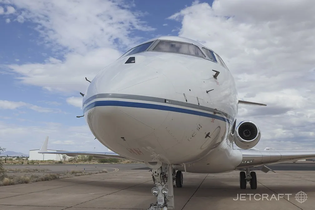 A close-up front view of the nose and cockpit of a white Bombardier Global 6000 business jet with blue and grey stripes.