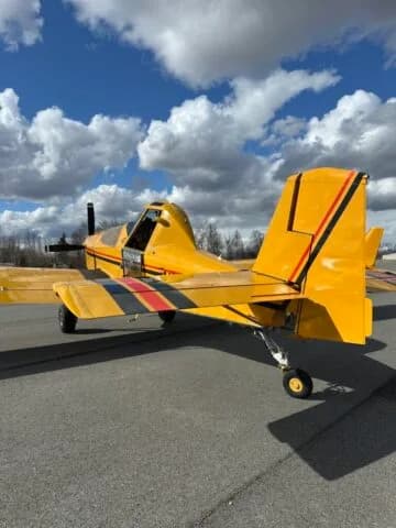 Rear three-quarter view of a yellow crop duster aircraft on an airfield under a cloudy blue sky.