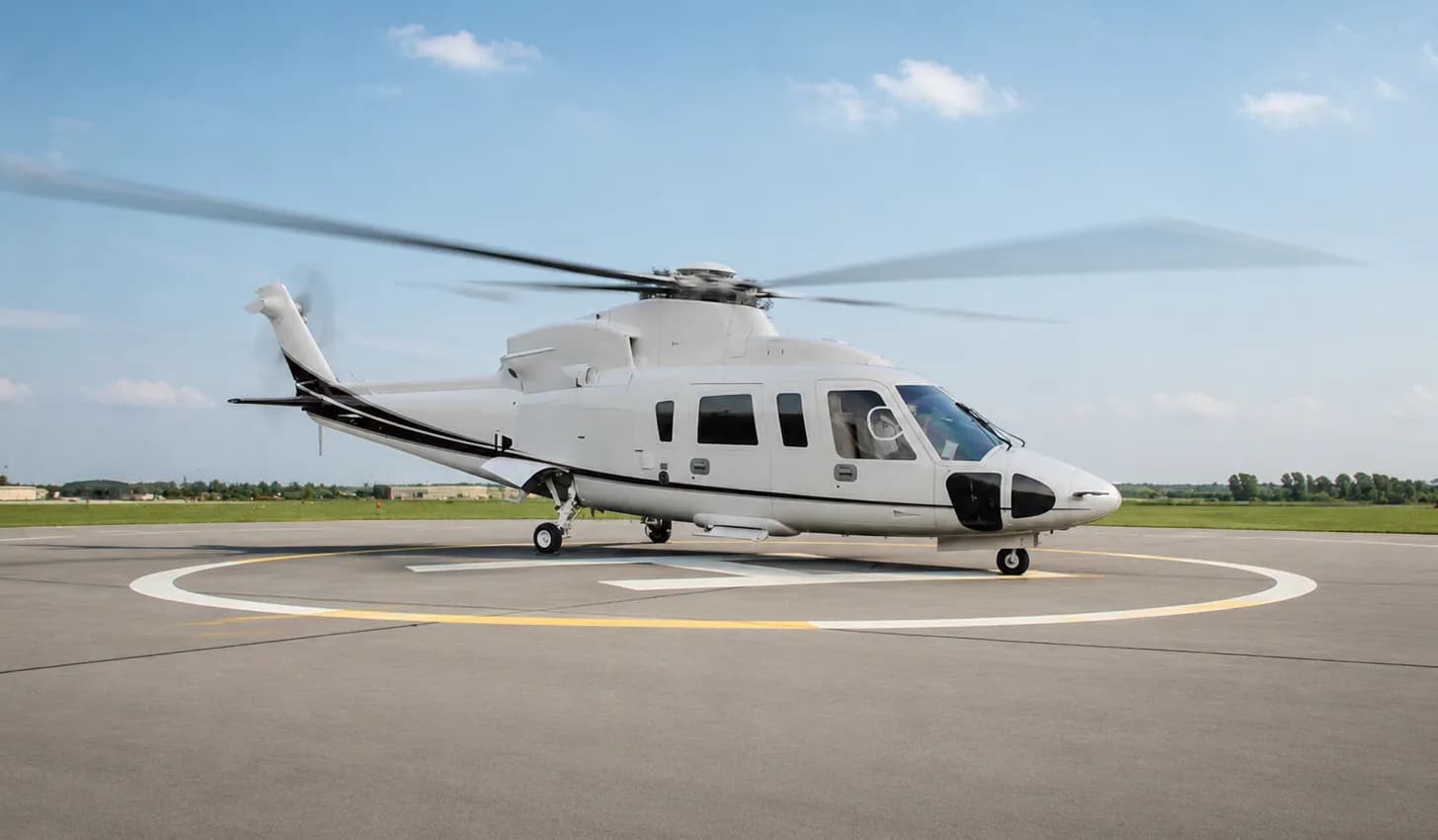 A white executive helicopter with black accents sits on a helipad with its rotors spinning under a blue sky.