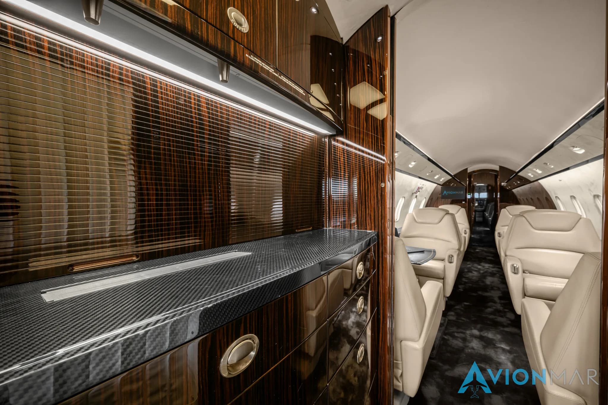 A view of the private jet's galley area with dark wood cabinetry and a carbon fiber countertop, looking towards the cabin.