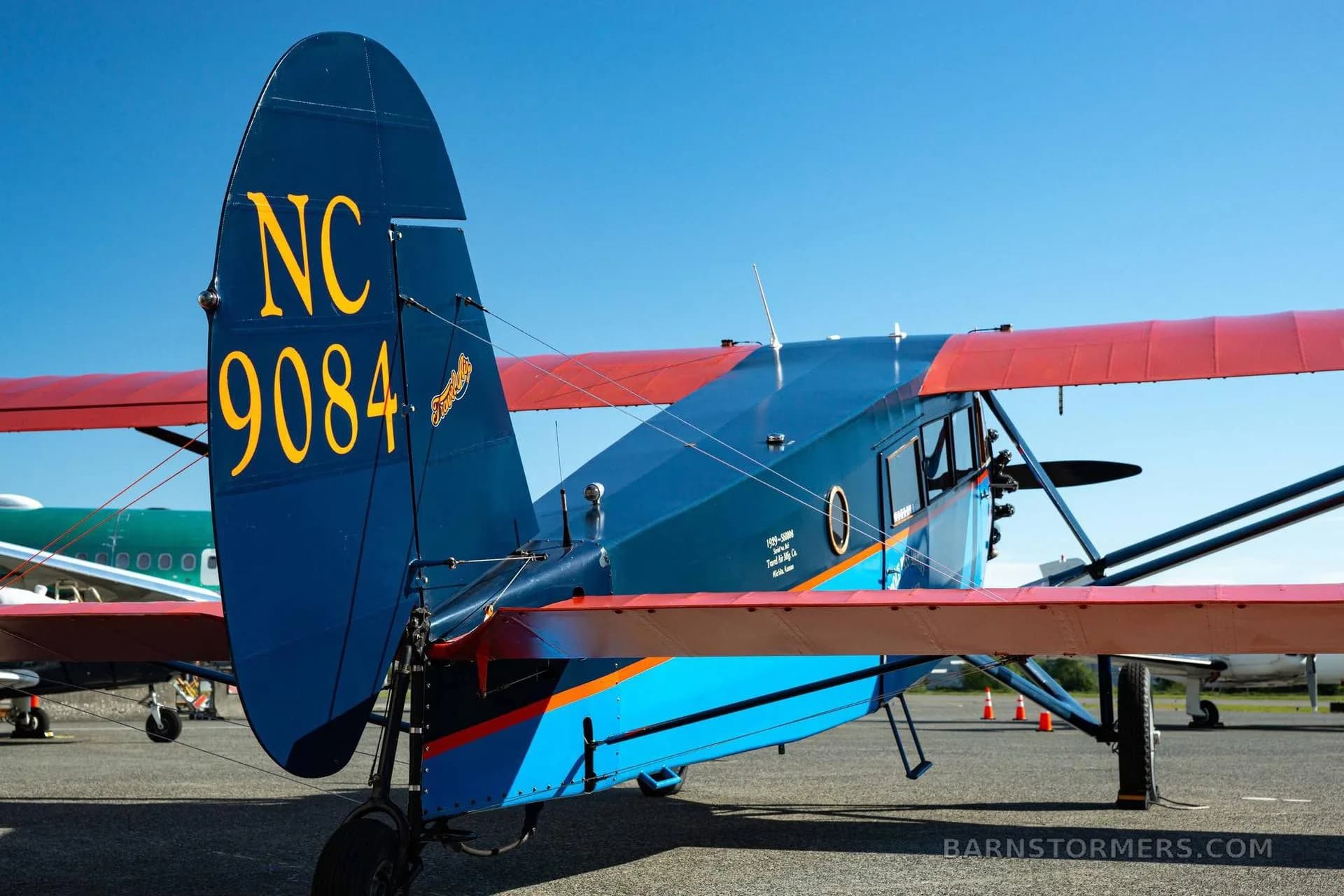 A rear view of a vintage blue and orange aircraft, showing its tail with registration NC 9084 and a partial view of another aircraft.