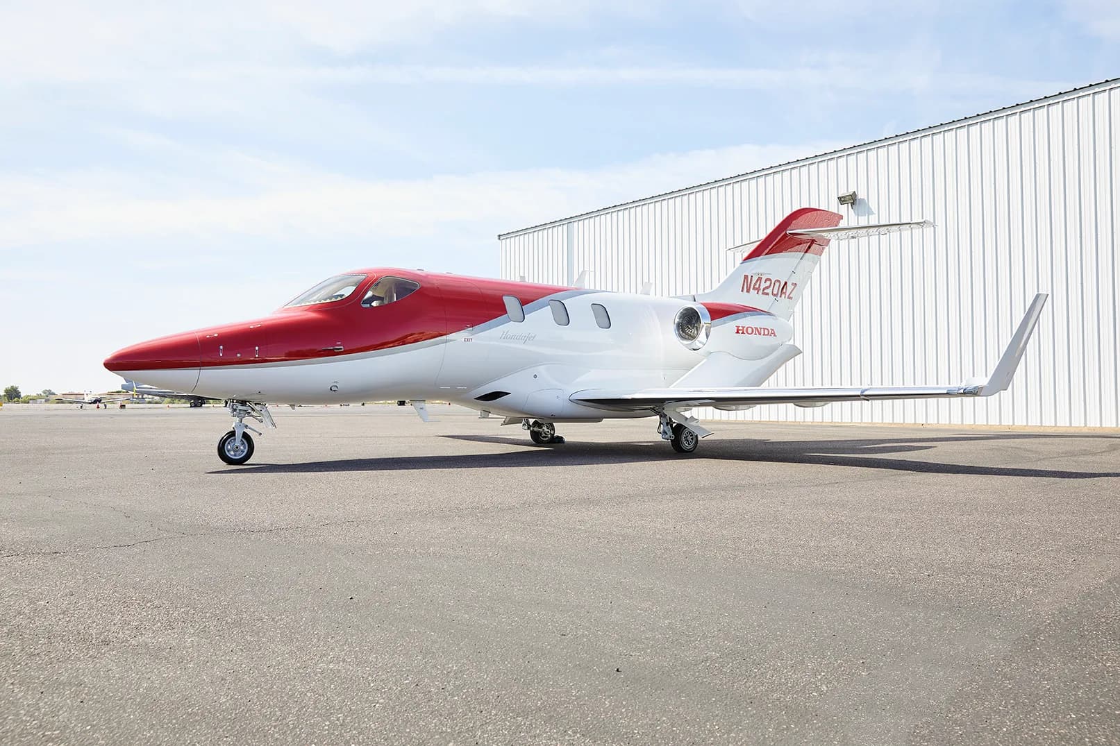 A red and white HondaJet HA-420 aircraft, registration N420AZ, is parked on an asphalt tarmac in front of a white hangar.