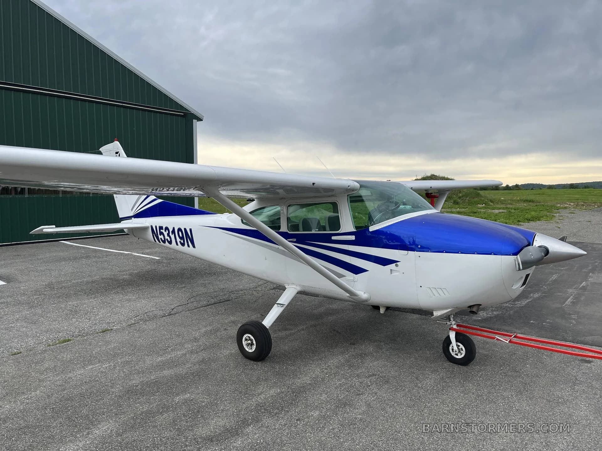 A white and blue Cessna aircraft with registration N5319N is parked on an asphalt ramp next to a green hangar under a cloudy sky.