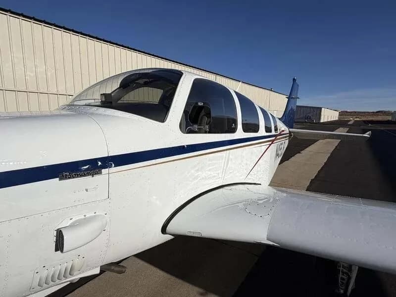 A side view of the cockpit and wing root of the white Beechcraft Bonanza, parked on an airfield.