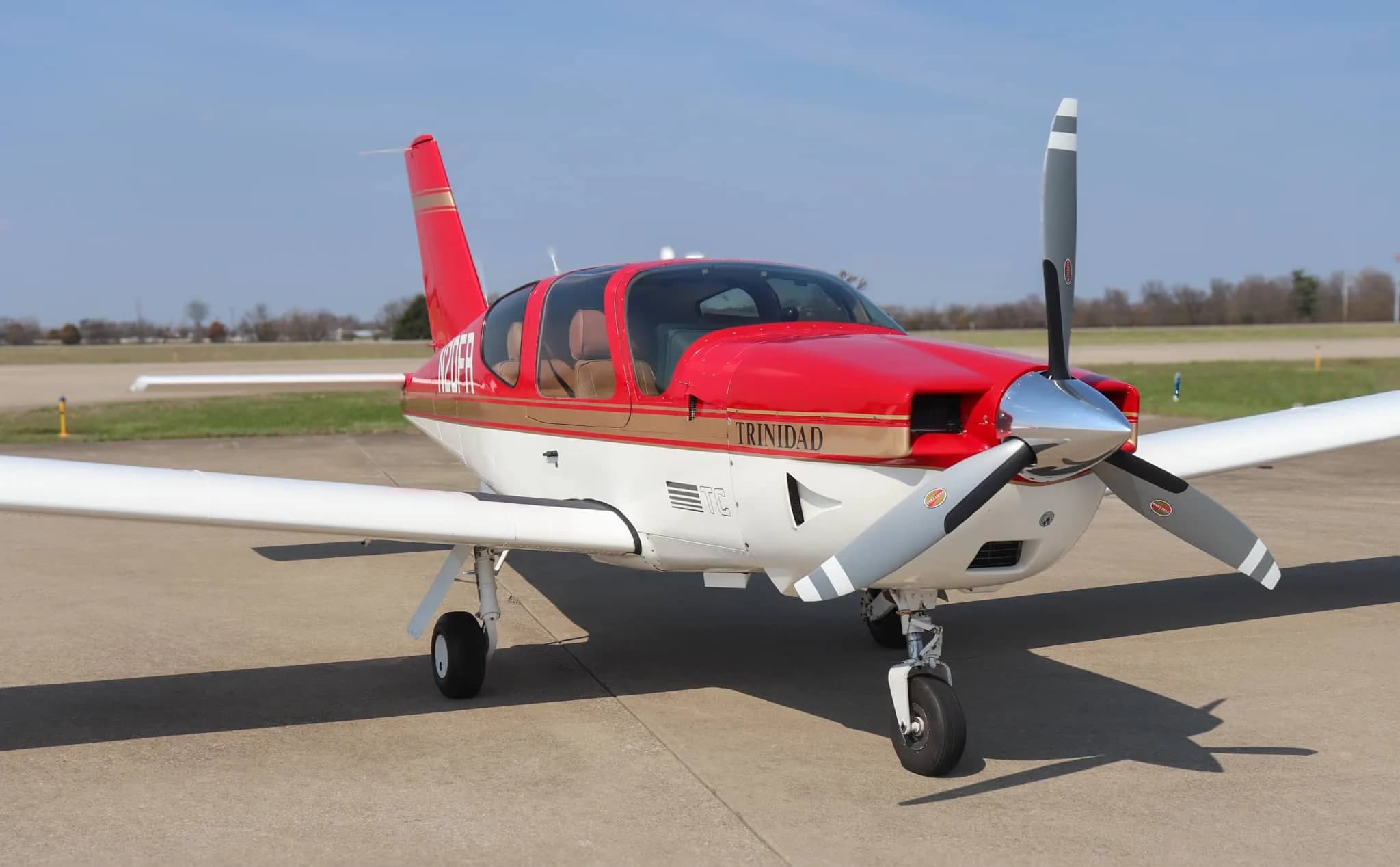 A red and white Socata Trinidad aircraft with gold stripes is parked on an airport tarmac, viewed from the front-left.
