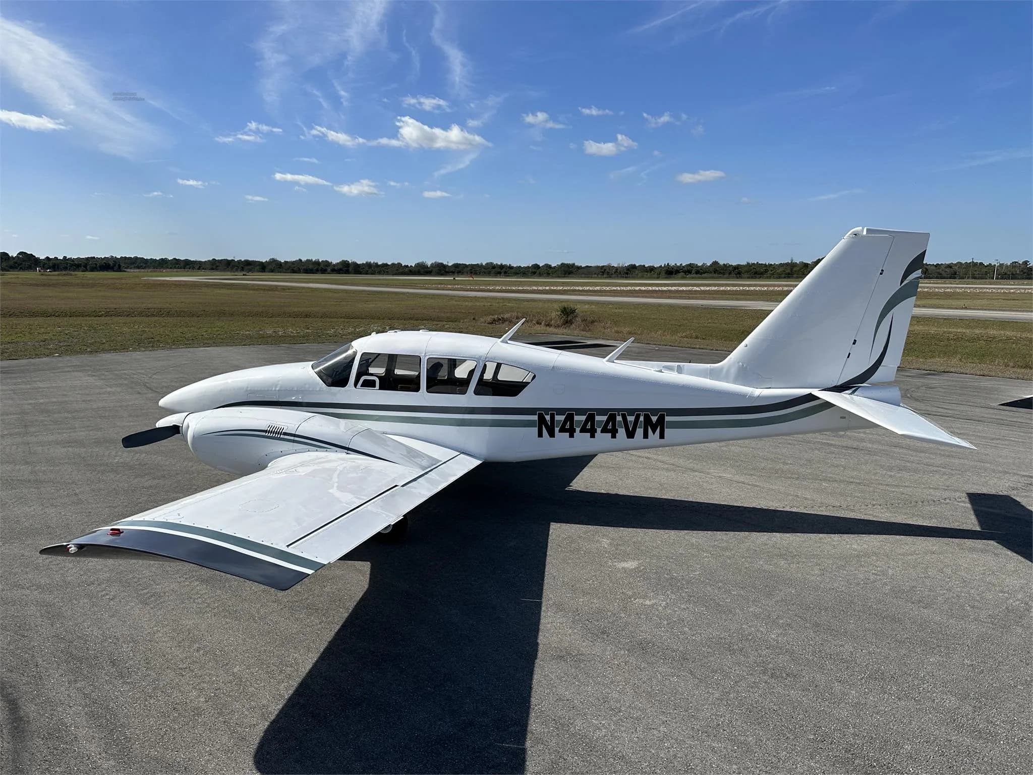 A rear-side view of a white Piper aircraft with green and black stripes, registration N444VM, parked on an airfield.
