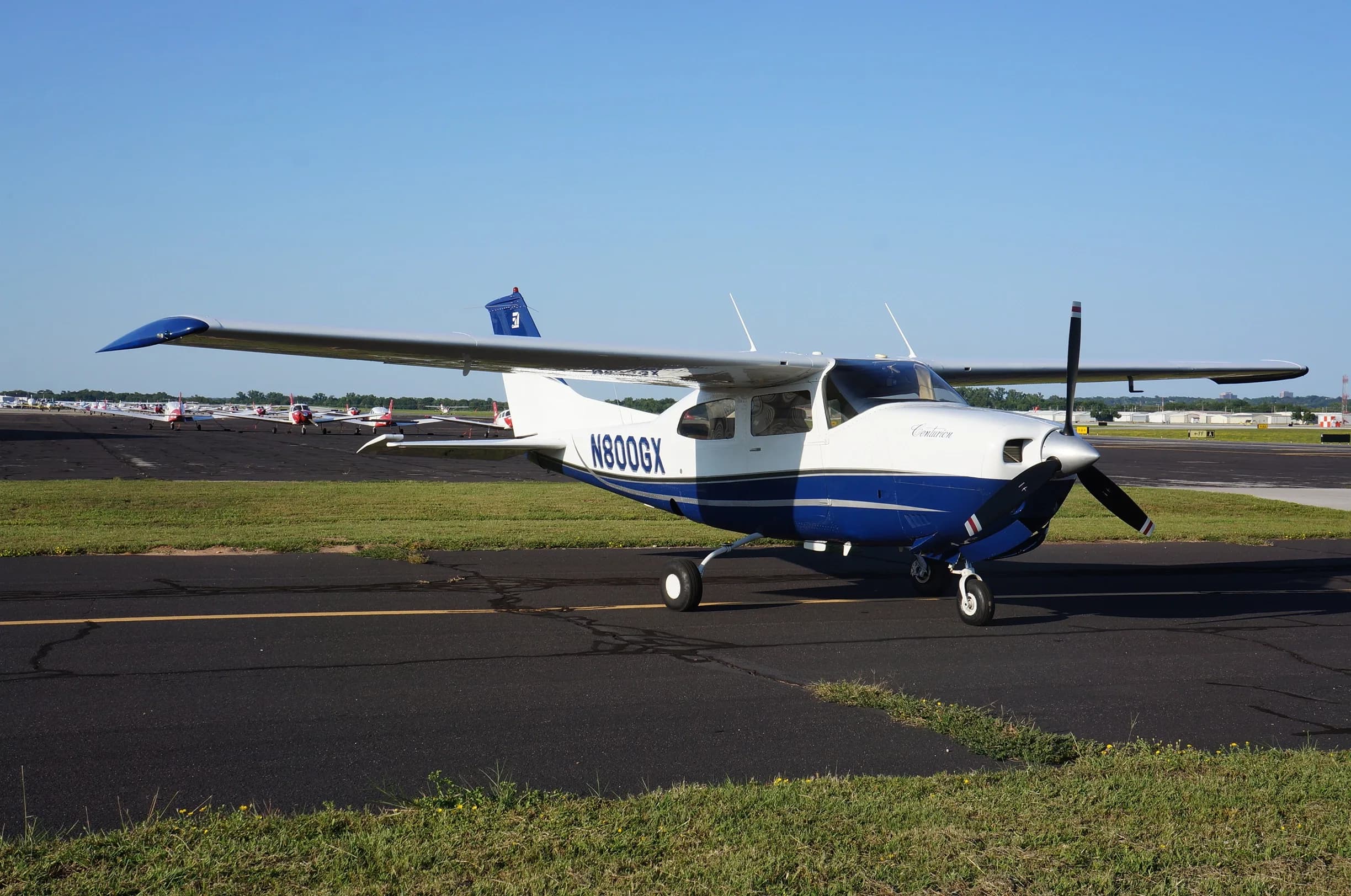A white and blue Cessna 206 Stationair aircraft, N800GX, is parked on an airport tarmac with other planes in the background.