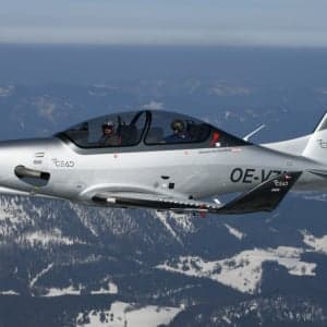 A silver CEAD aircraft flies over a snowy mountain range.