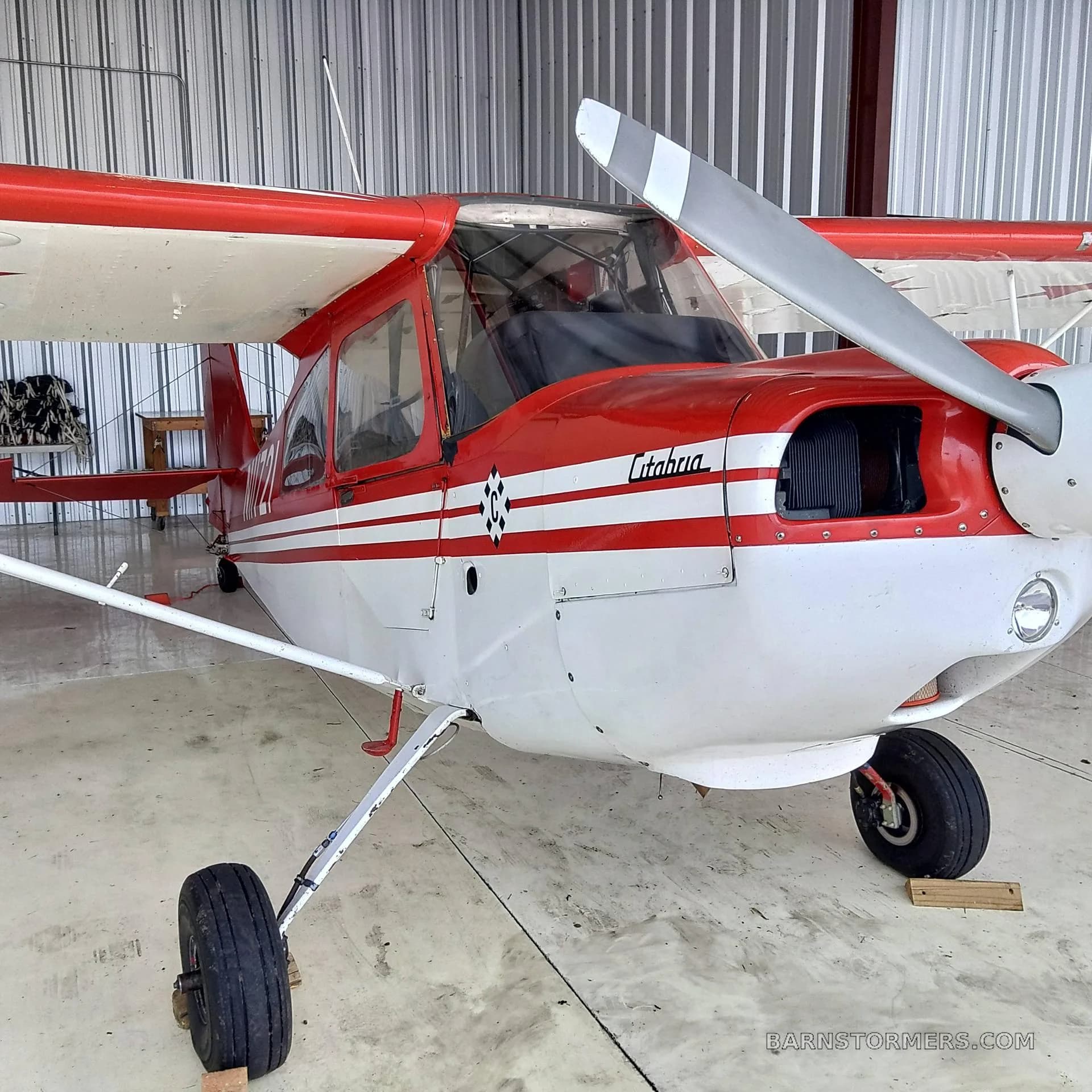 A close-up front view of a red and white Citabria N11721 aircraft with a propeller, parked in a hangar.