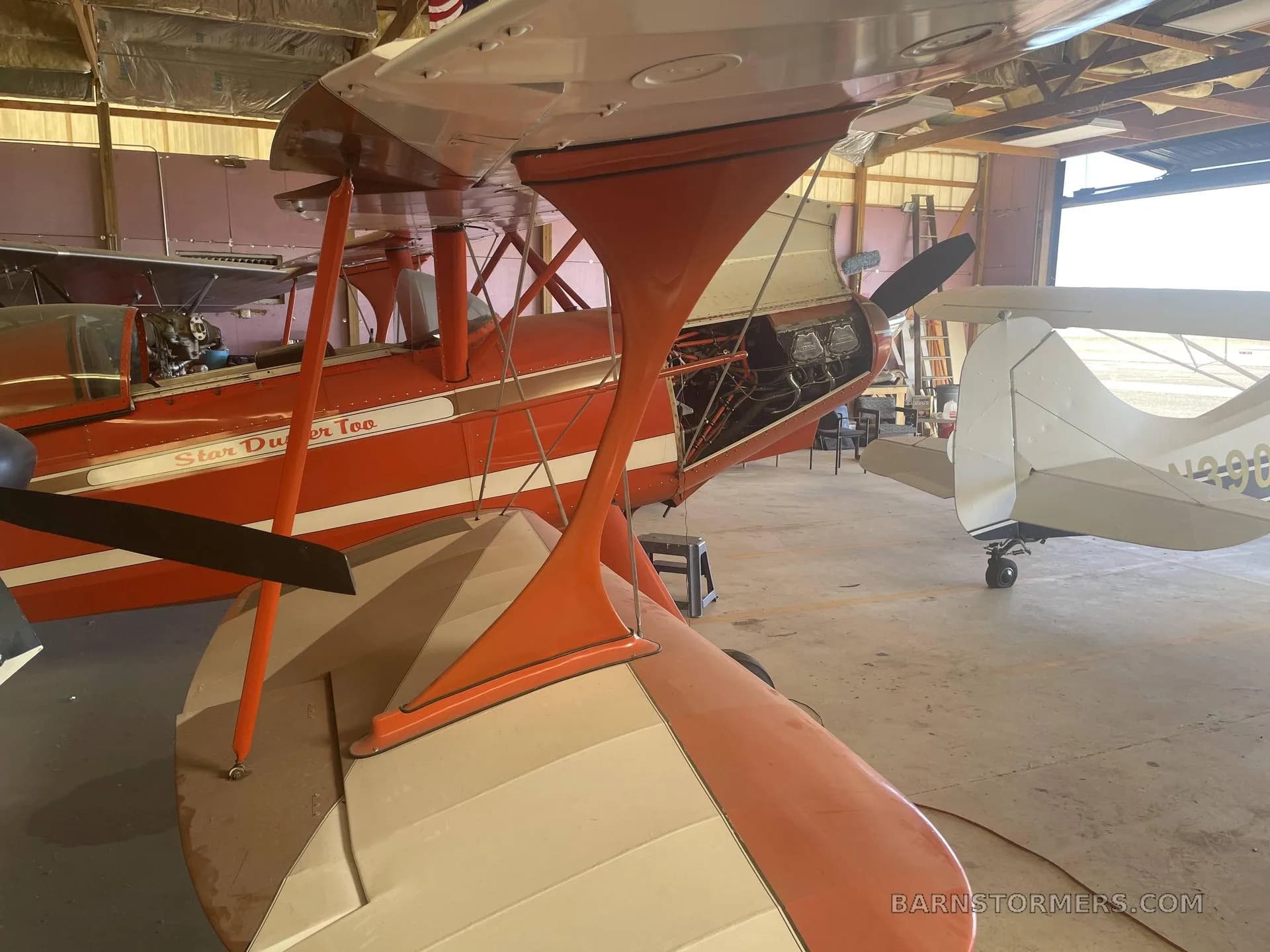 An orange and white Star Duster Too biplane with its engine cowling open is parked in a hangar next to another small aircraft.