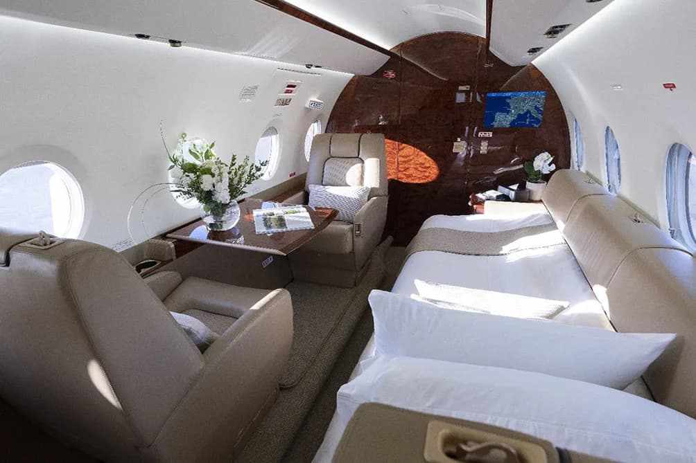 A view of the private jet's cabin, showing a bed, a table with a floral arrangement, and beige seating.