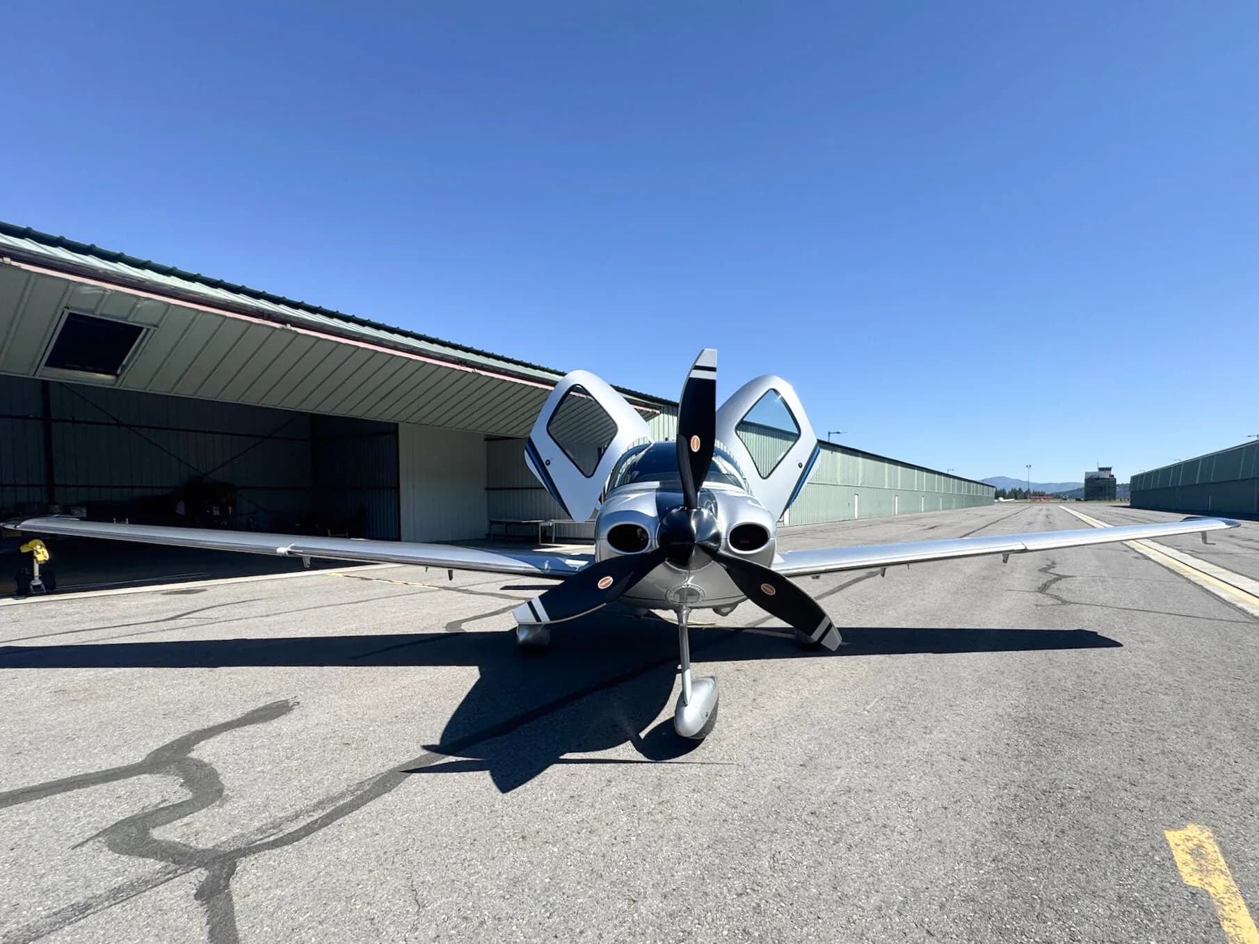 A front view of a silver and black Cirrus SR22T aircraft with N191WL, parked on an asphalt apron with both doors open, facing a hangar.