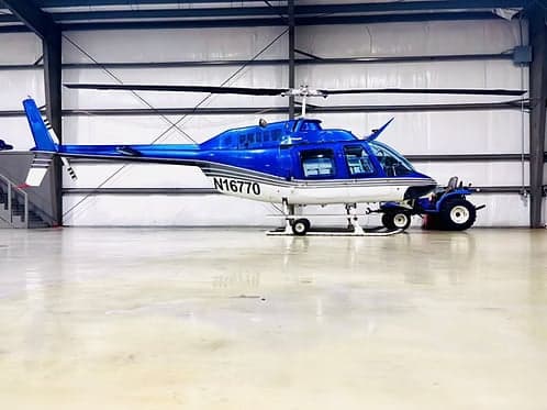 A blue and white Bell 407 helicopter, registration N16770, parked in a hangar with a tractor attached.