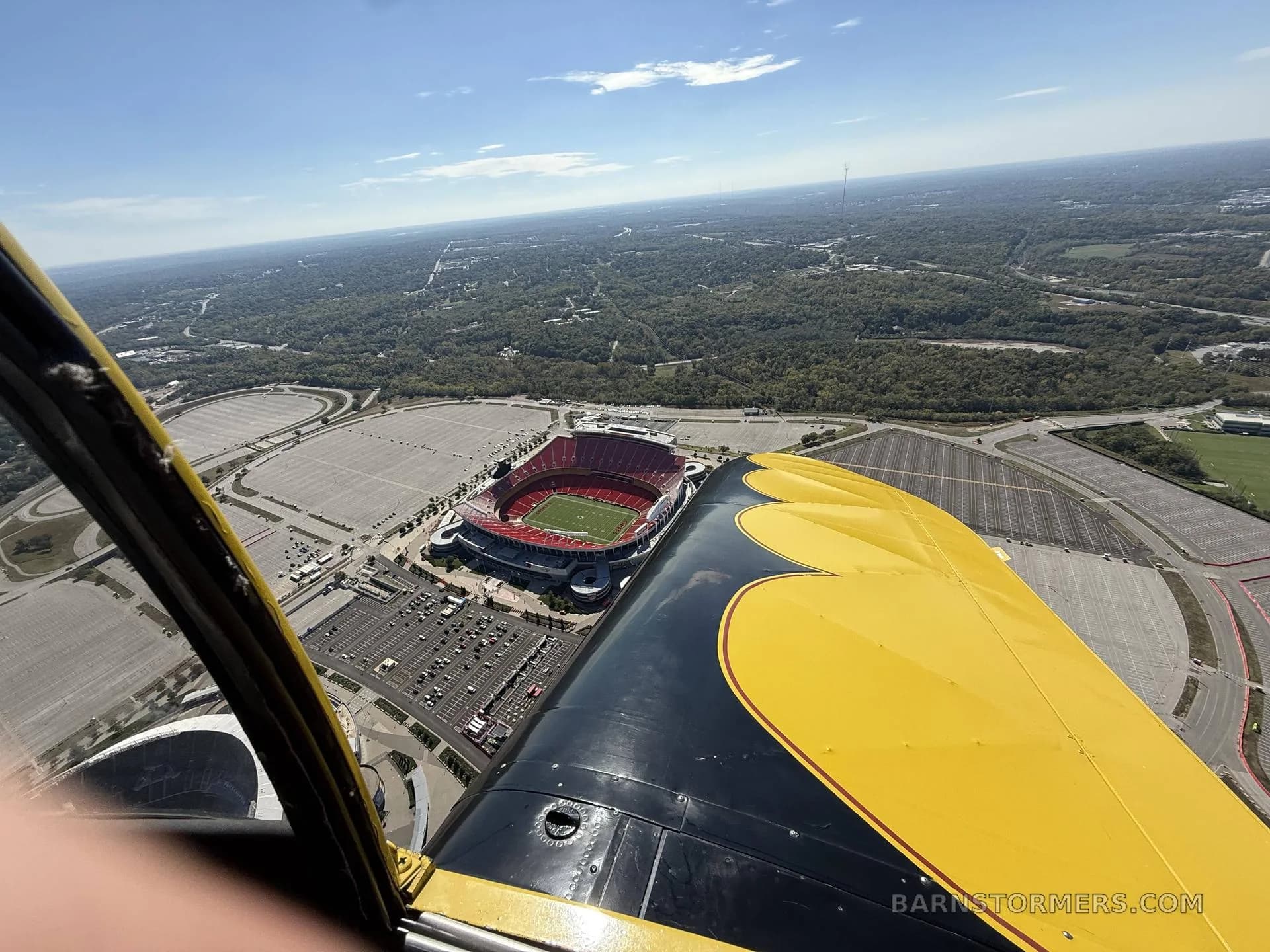 An aerial view from a black and yellow aircraft, showing its wing and a large sports stadium with surrounding parking lots below.