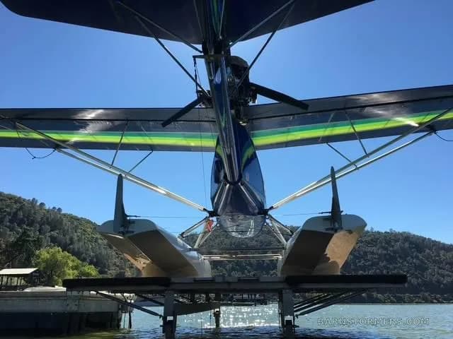 A low-angle view from behind a blue and green seaplane showing its tail, propeller, and floats docked on a lake.