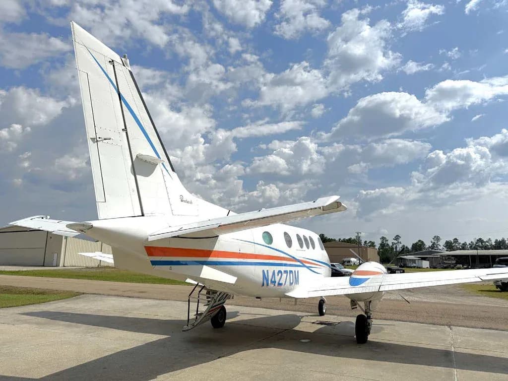 The rear view of a white twin-engine turboprop aircraft with blue and orange stripes, registration N427DM, parked on a tarmac.