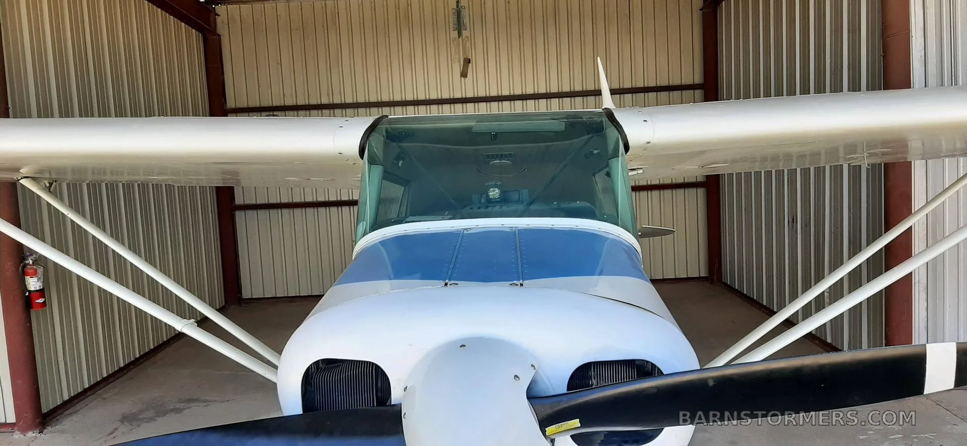 A front view of a white and blue single-engine aircraft with a propeller, parked inside a hangar.