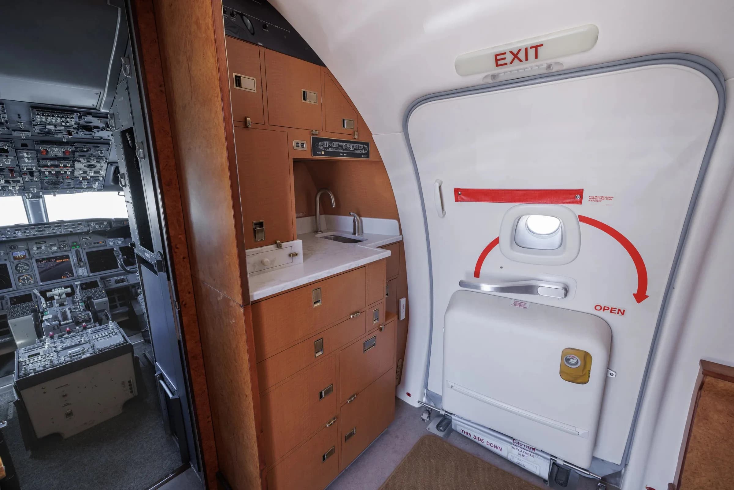 The aircraft's galley area with wooden cabinetry, a sink, and the main entrance door, with the cockpit visible through an open doorway.
