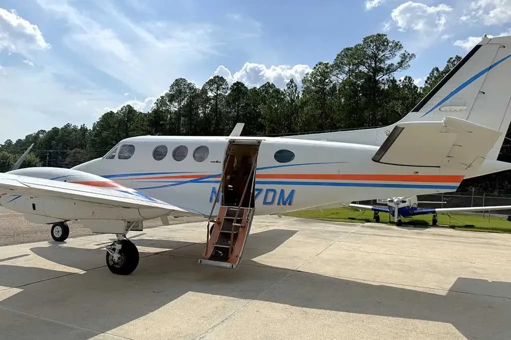 A white twin-engine turboprop aircraft with blue and orange stripes, registration N427DM, parked on a tarmac with its passenger door open.
